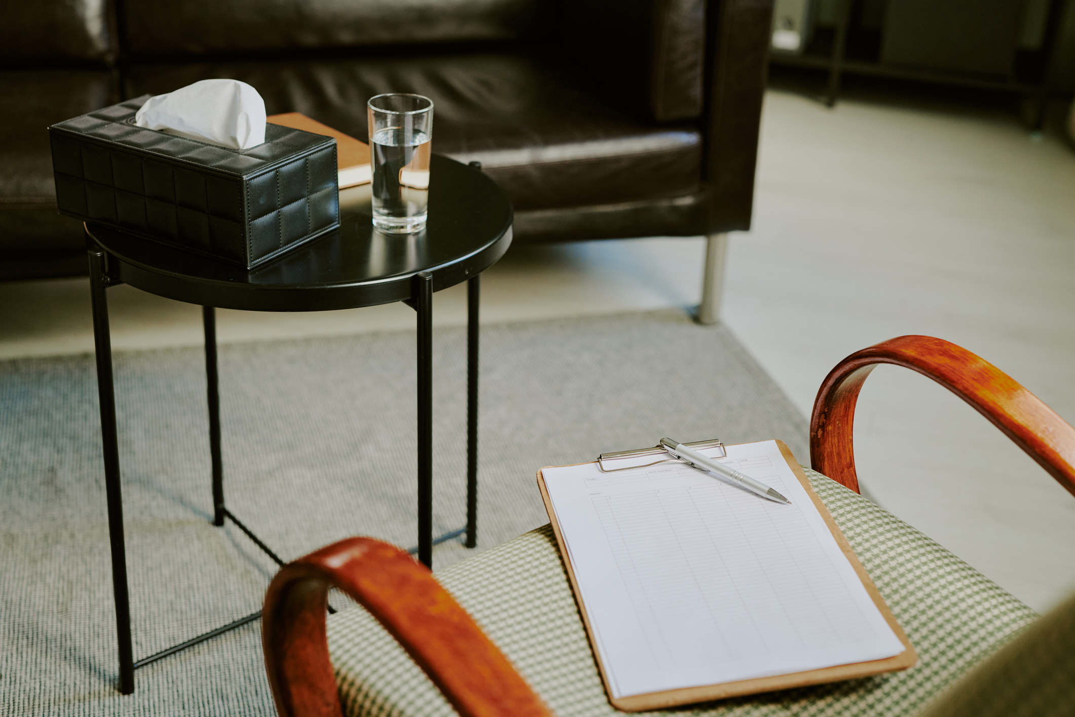 A cozy counselling room with a round black table holding a tissue box, a glass of water, and a notebook nearby. A clipboard and a pen are on an armchair.