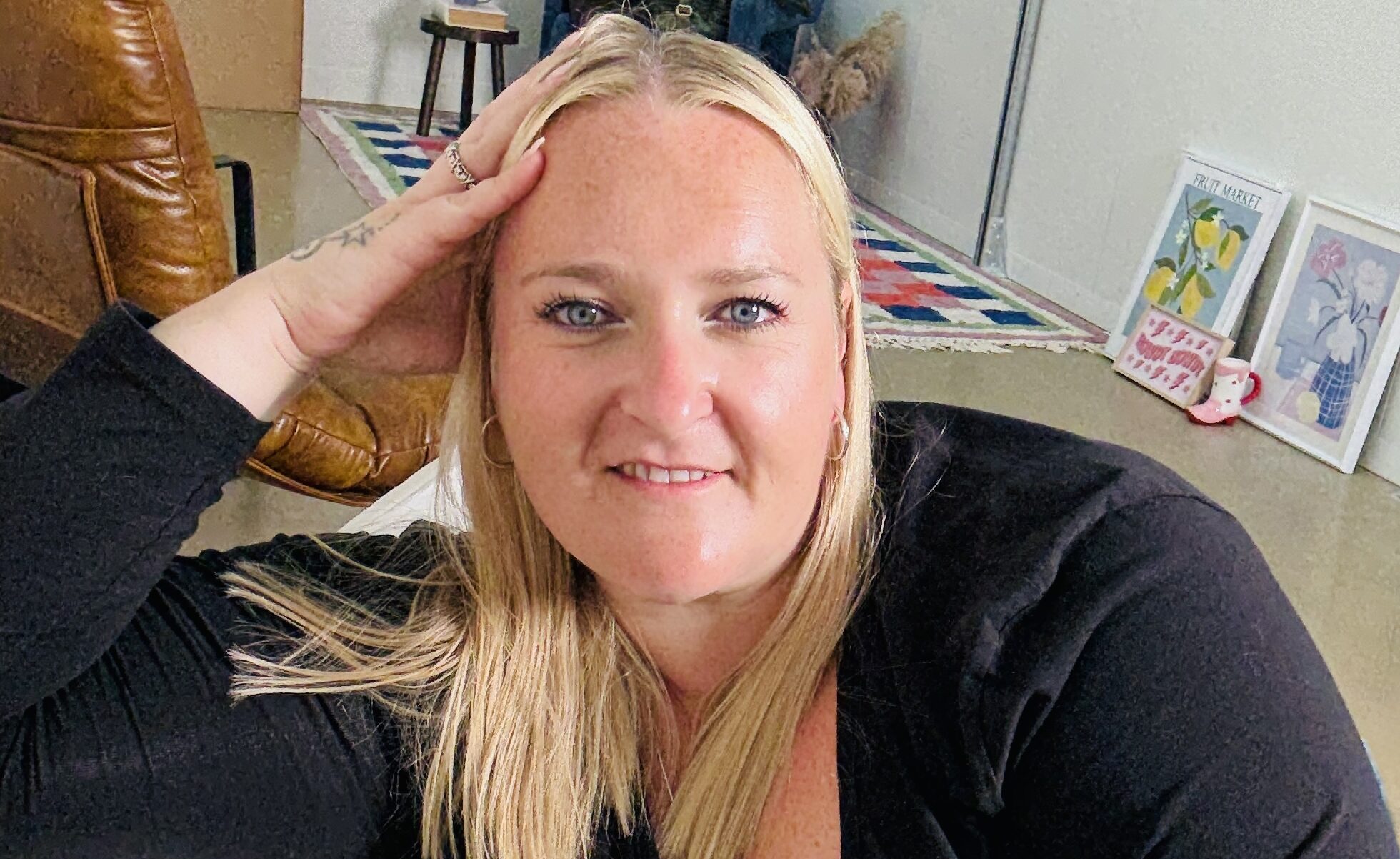 A zoomed-in picture of Kerry Pitt wearing a black long-sleeved top smiles warmly as she rests her head on her hand and looks directly at the camera. Behind her, the space is filled with colourful framed artwork and decorative accents, with a brown leather chair and a patterned rug.