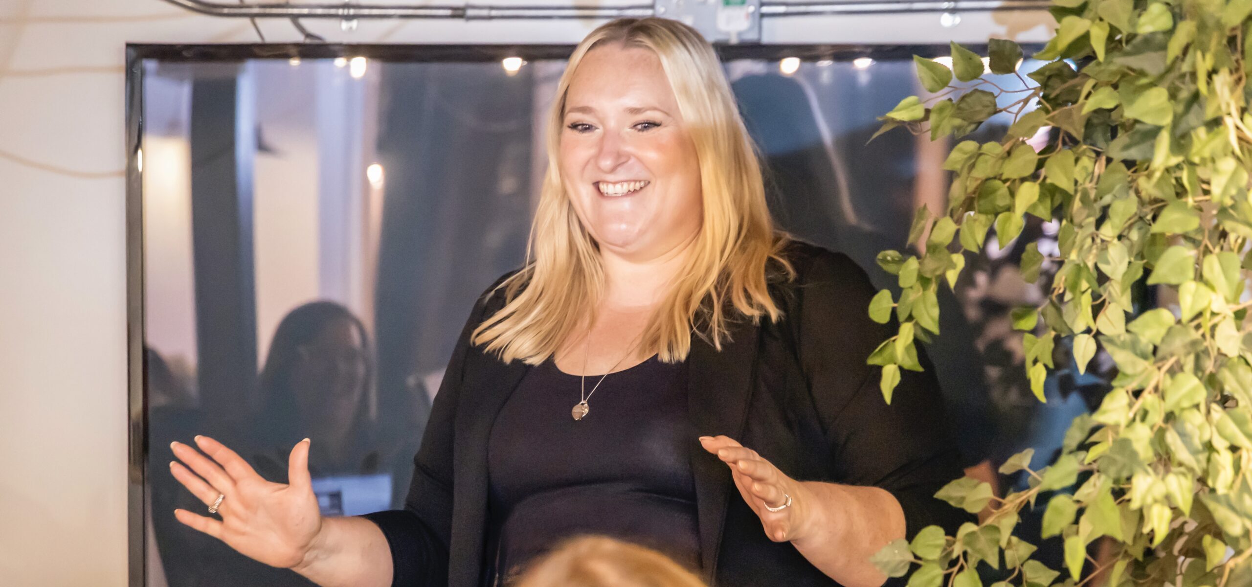 A zoomed-in picture of Kerry Pitt in a black blazer speaks and gestures to an audience, with a leafy plant and a dark screen in the background, where the audience is faintly reflected.
