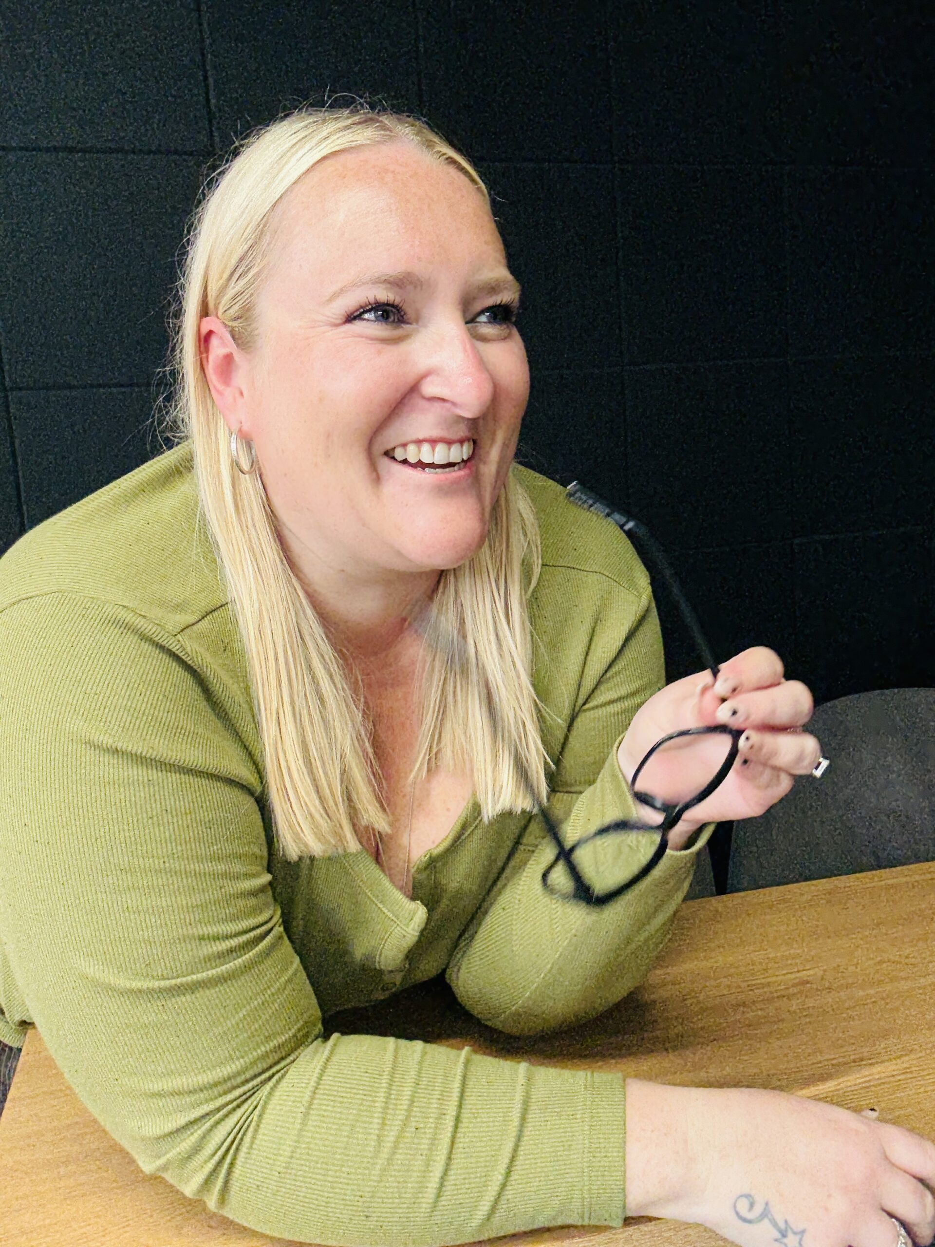 Kerry Pitt wears an olive green top and sits, holding her glasses in front of her against a dark backdrop. She is smiling and looking towards the right.