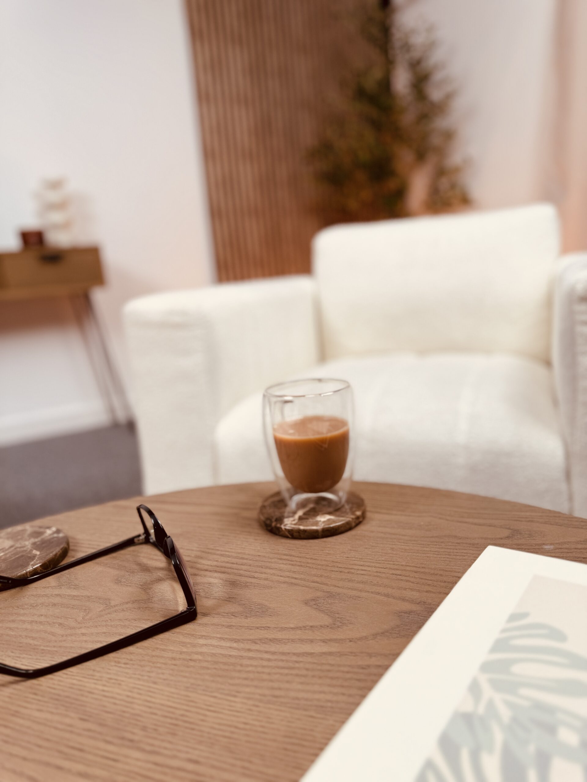 A warm living room scene with a white armchair and wooden table in the foreground holding a glass of coffee and glasses, set against a background of wooden panelling, a white wall, a plant, and a wooden console desk.