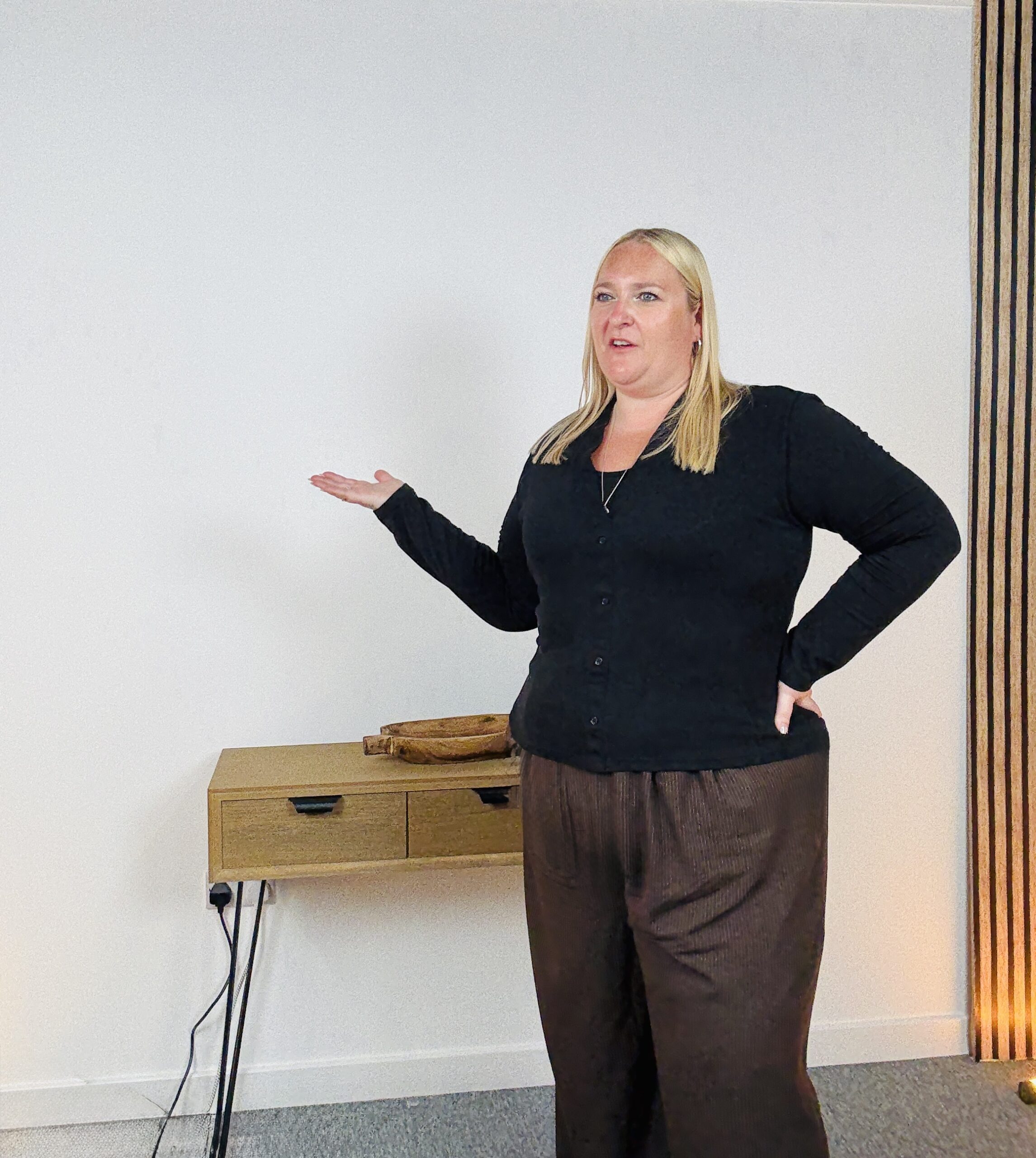 Kerry Pitt stands in a neutral-toned room, gesturing with one hand as she is captured mid-sentence. She wears a black cardigan and brown trousers, with a wooden console table behind her.