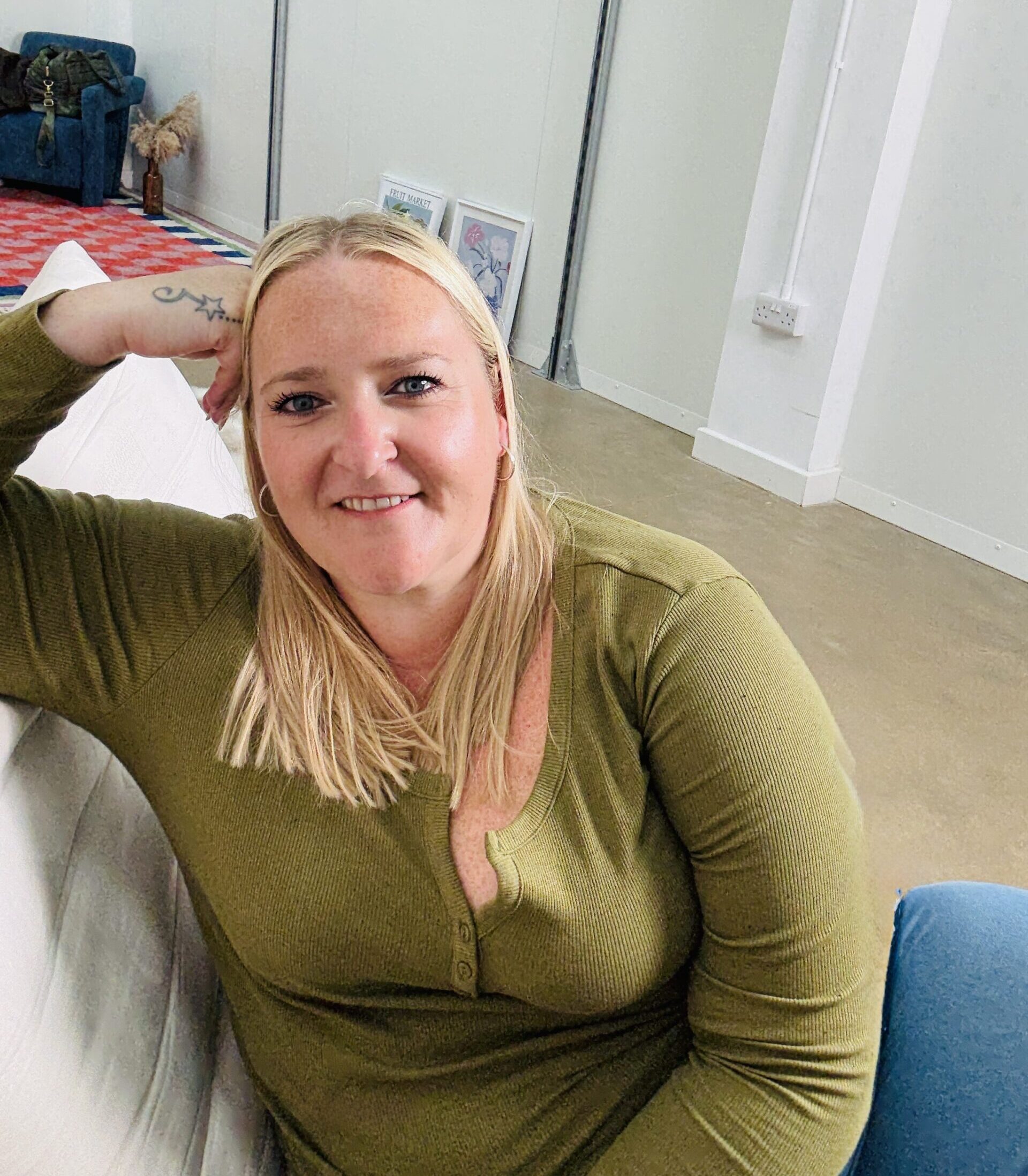 Kerry Pitt sits on a cream-colored chair, smiling at the camera while leaning her head on her hand. Behind her, a mirror reflects part of the room, with a patterned rug, a blue armchair, 2 famed prints and a plant.